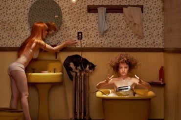 Two women appear in a square cropped, stylised image. One woman stands at the bathroom sink whilst another appears in the bathtub who has accidentally dropped a toaster into the water and she is surprised and shocked at her mistake.